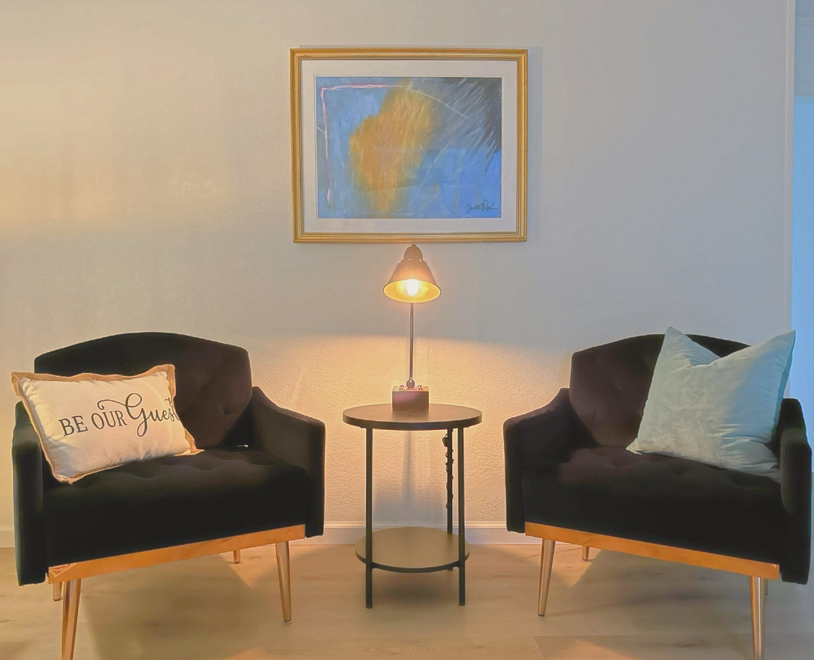 Two velvet chairs with a brass lamp and a gold-framed painting between them