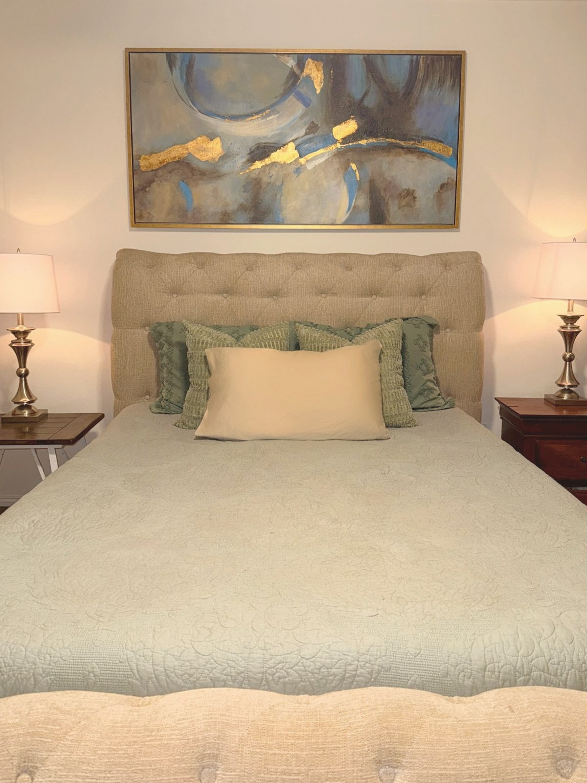 Primary bedroom with a tufted headboard, sage pillows, brass lamps, and a gold-framed painting