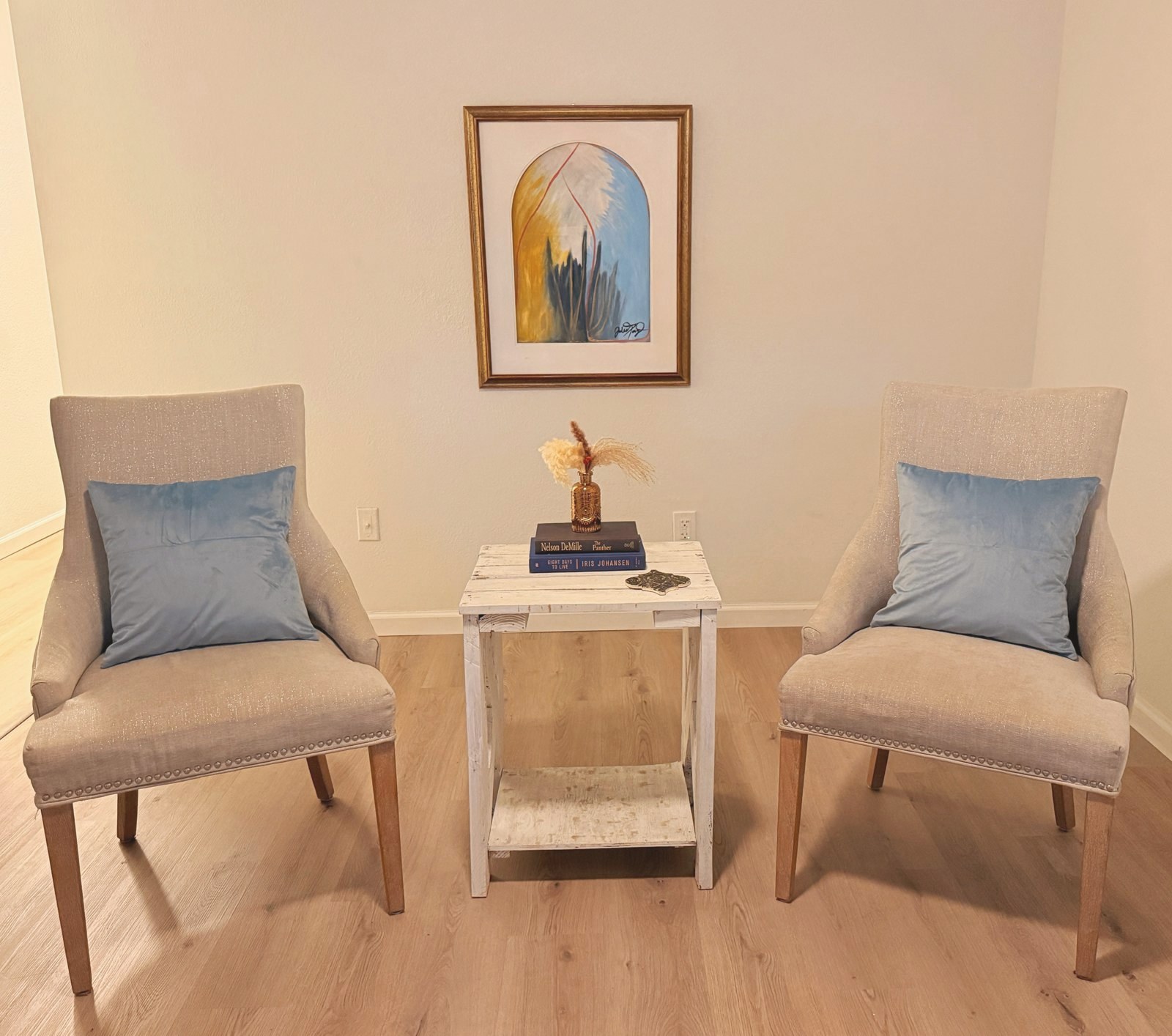 Two linen chairs with blue pillows and dried grass on a white side table