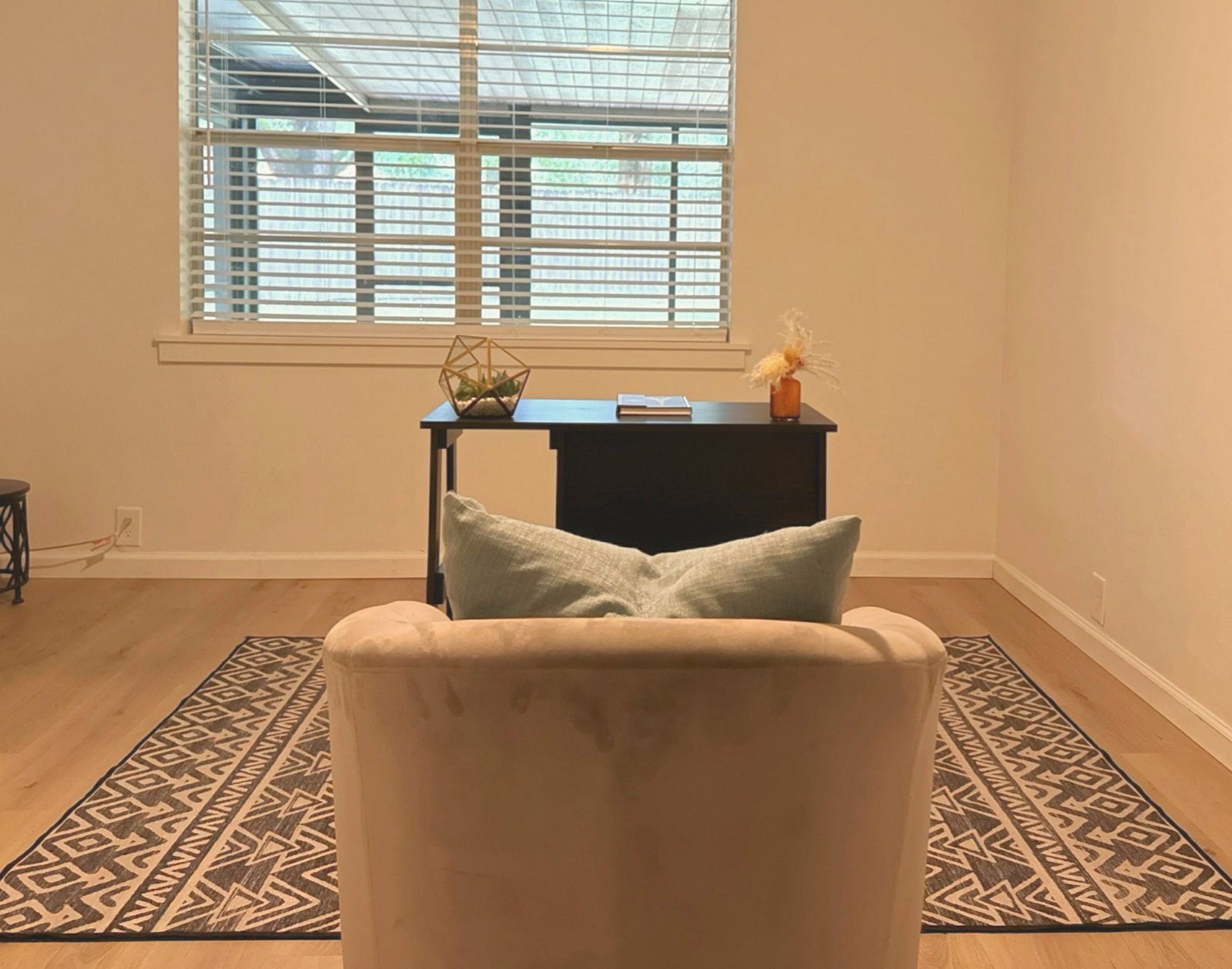 View from a blush chair toward a desk styled with a book, terrarium, and pampas