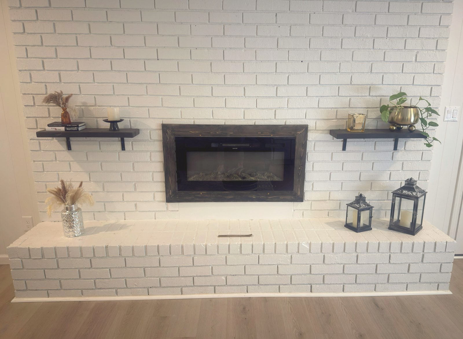 White brick fireplace styled with shelves, lanterns, candles, and pampas grass
