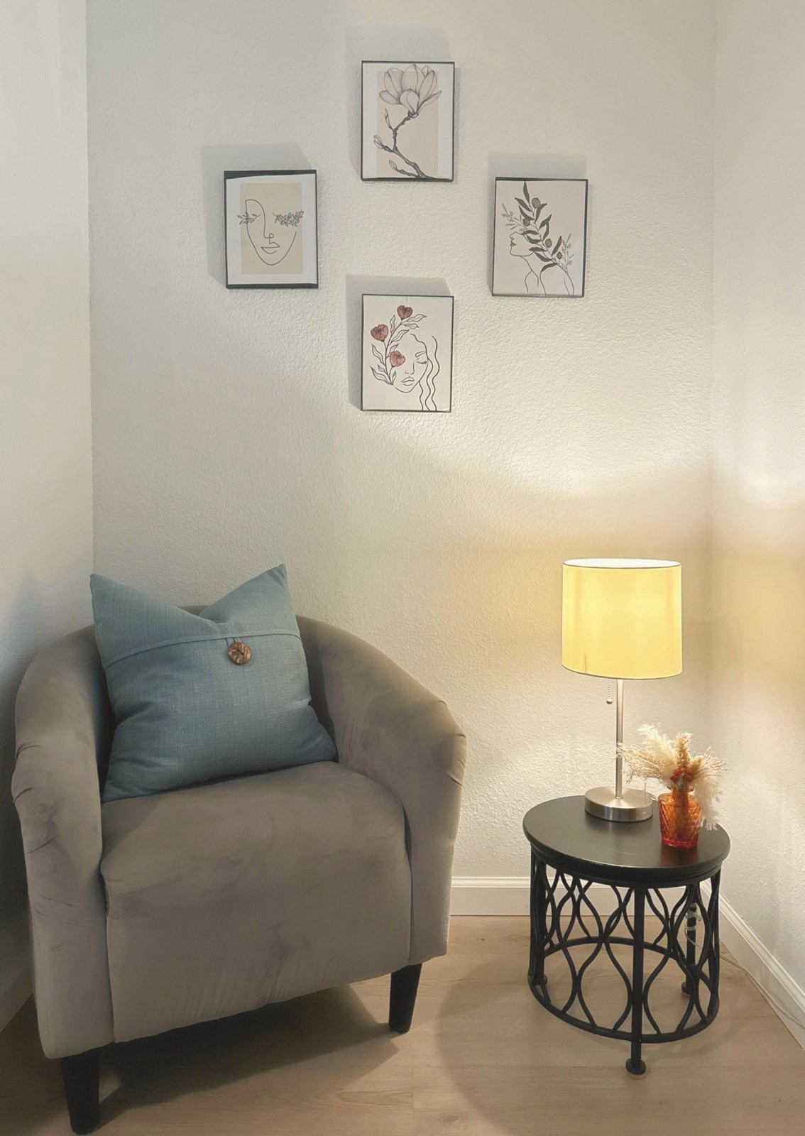 Gray chair with a blue pillow beside a lamp and four framed line drawings