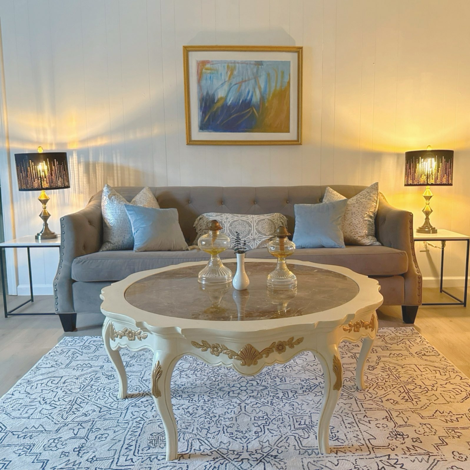 Gray tufted sofa with blue pillows, brass lamps, and a marble-top coffee table